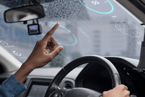 A person inside a car interacts with a futuristic augmented reality dashboard projected onto the windshield, featuring various digital gauges and information. The driver monitoring system enhances safety as the drivers hand reaches towards the virtual interface.