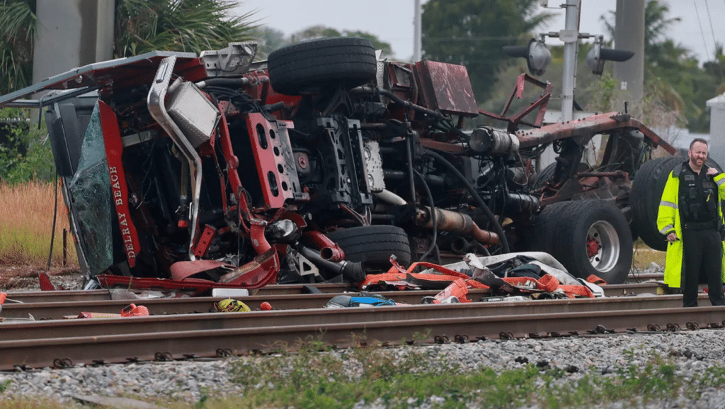 A red tow truck is flipped on its side, resting across train tracks—a stark reminder of work-related car accidents. Debris is scattered around as a uniformed person surveys the scene. In the background, trees and a utility pole stand under an overcast sky.