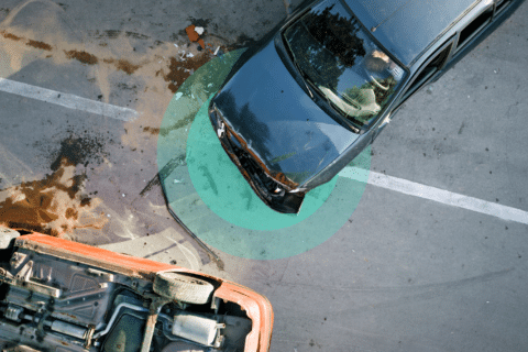 Aerial view of a work-related car accident involving an overturned orange car and a black car with a damaged front. The vehicles are on a street marked by visible skid marks and fluid spill, highlighted by a green circular overlay.