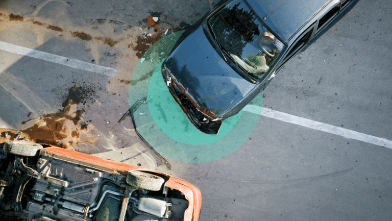 Aerial view of a work-related car accident involving an overturned orange car and a black car with a damaged front. The vehicles are on a street marked by visible skid marks and fluid spill, highlighted by a green circular overlay.