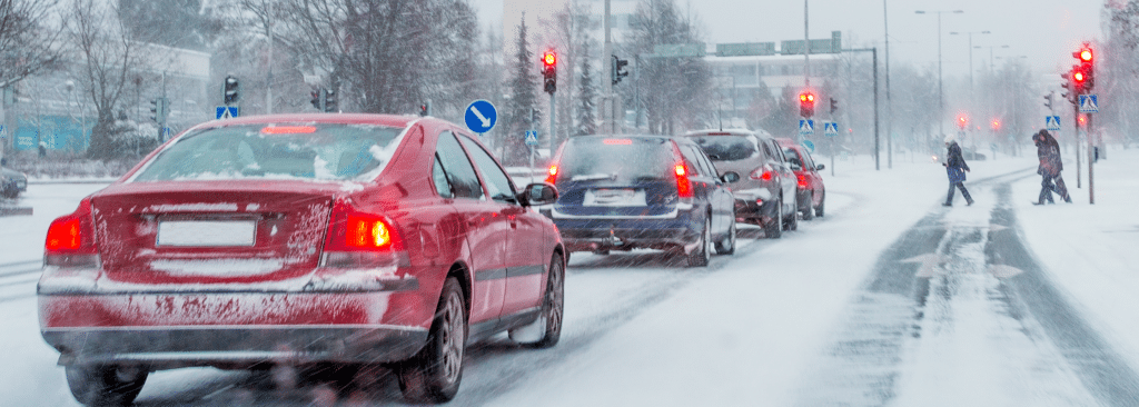 Cars lined up at a snowy intersection with red traffic lights. The road is covered in snow, and pedestrians are crossing. Trees and buildings are in the background under overcast skies.