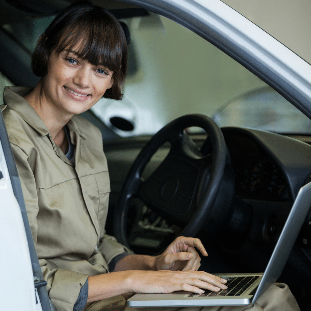 A woman in a mechanics uniform sits in a car with an open door, smiling and typing on a laptop. With the steering wheel and car interior visible against the garage backdrop, it seems shes integrating Fleet Driver Training insights into her work.