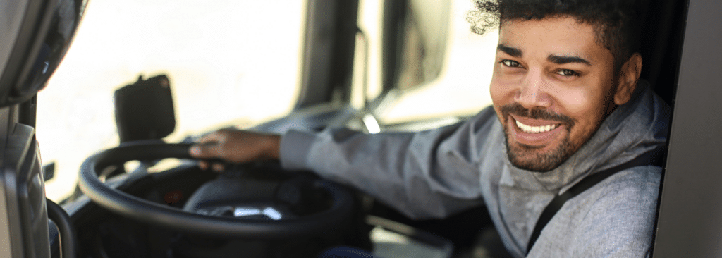 Man with curly hair and a beard sitting in a vehicle, smiling at the camera. He is wearing a gray sweatshirt and gripping the steering wheel, suggesting he is the driver. The background is blurred, focusing on his cheerful expression.