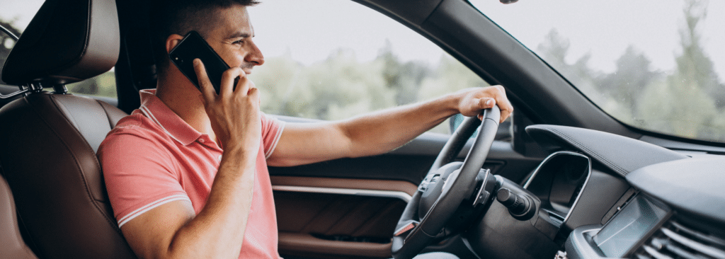 Man driving a car while talking on a smartphone. He is wearing a pink polo shirt and is holding the steering wheel with his left hand. The car interior is modern with a visible dashboard and a blurred green outdoor background.