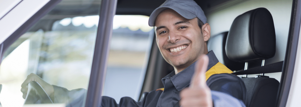 A person with a cap is smiling and giving a thumbs-up gesture while sitting in the drivers seat of a vehicle, looking towards the camera. The background is slightly blurred.