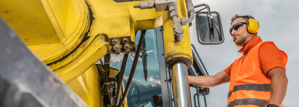 A construction worker wearing an orange safety vest and headphones operates heavy machinery. The machinery is yellow with metallic components. The worker is focused and wearing sunglasses. The sky is visible in the background.